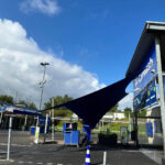 Outdoor car wash facility with a dark blue shade sail, blue signage, and service bays under a bright blue sky.