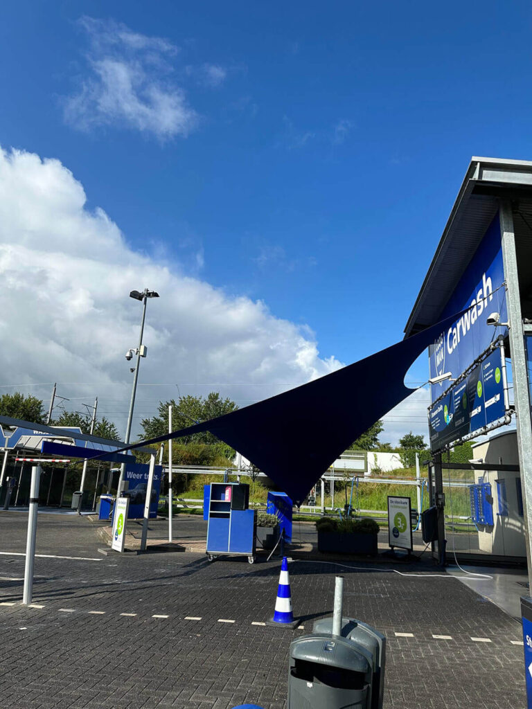 Outdoor car wash facility with a dark blue shade sail, blue signage, and service bays under a bright blue sky.