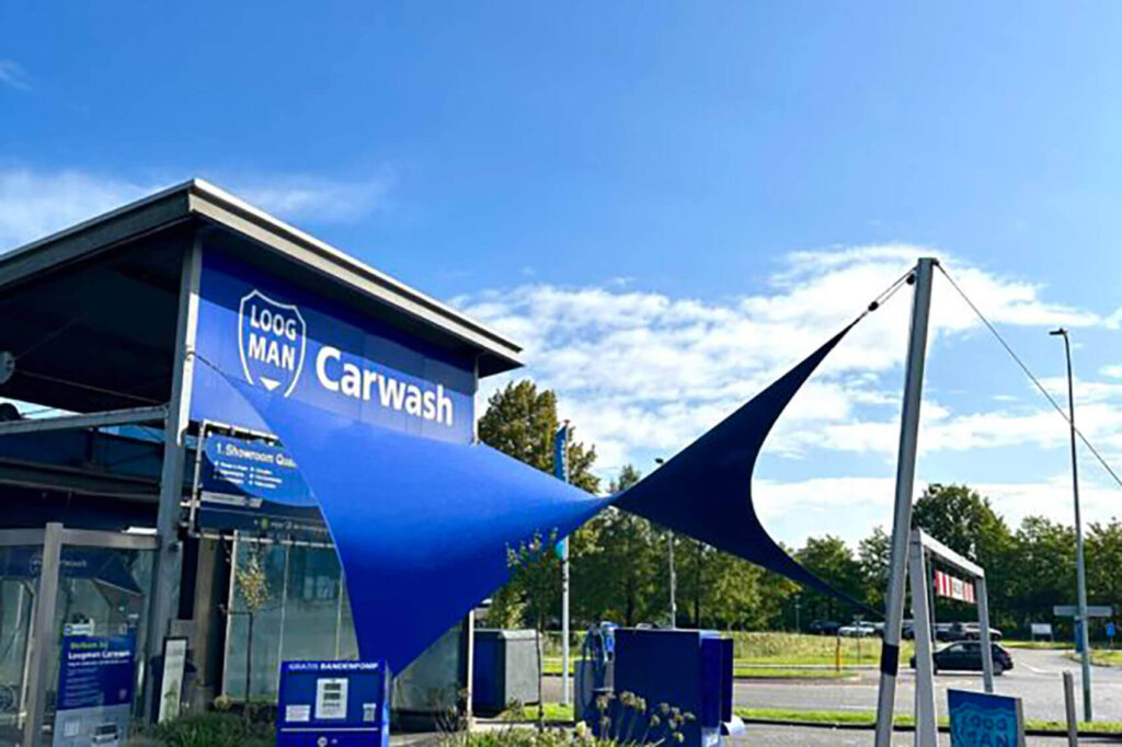 Car wash building with blue signage reading 'Carwash' and blue shade sails in front under a clear sky.