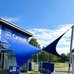 Car wash building with blue signage reading 'Carwash' and blue shade sails in front under a clear sky.