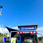 Car wash facility with a silver SUV entering; large blue sign reading 'Carwash' above the bay and a height limit bar across the entrance.