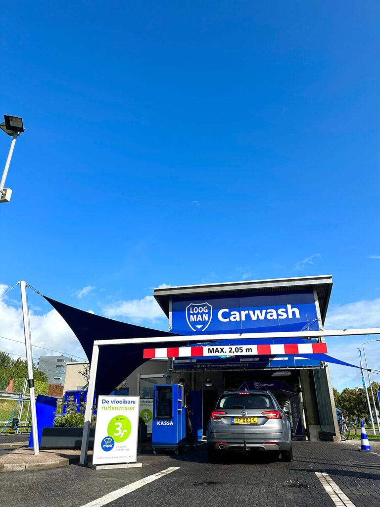 Car wash facility with a silver SUV entering; large blue sign reading 'Carwash' above the bay and a height limit bar across the entrance.