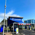 Carwash building with a blue sign that reads 'Carwash' under a bright blue sky, with a dark blue sail shade extending over the entrance area.