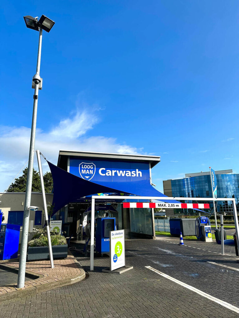 Carwash building with a blue sign that reads 'Carwash' under a bright blue sky, with a dark blue sail shade extending over the entrance area.