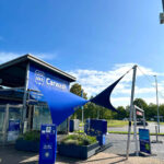 Exterior view of a carwash with blue signage and large fabric shade sails over the entrance area.