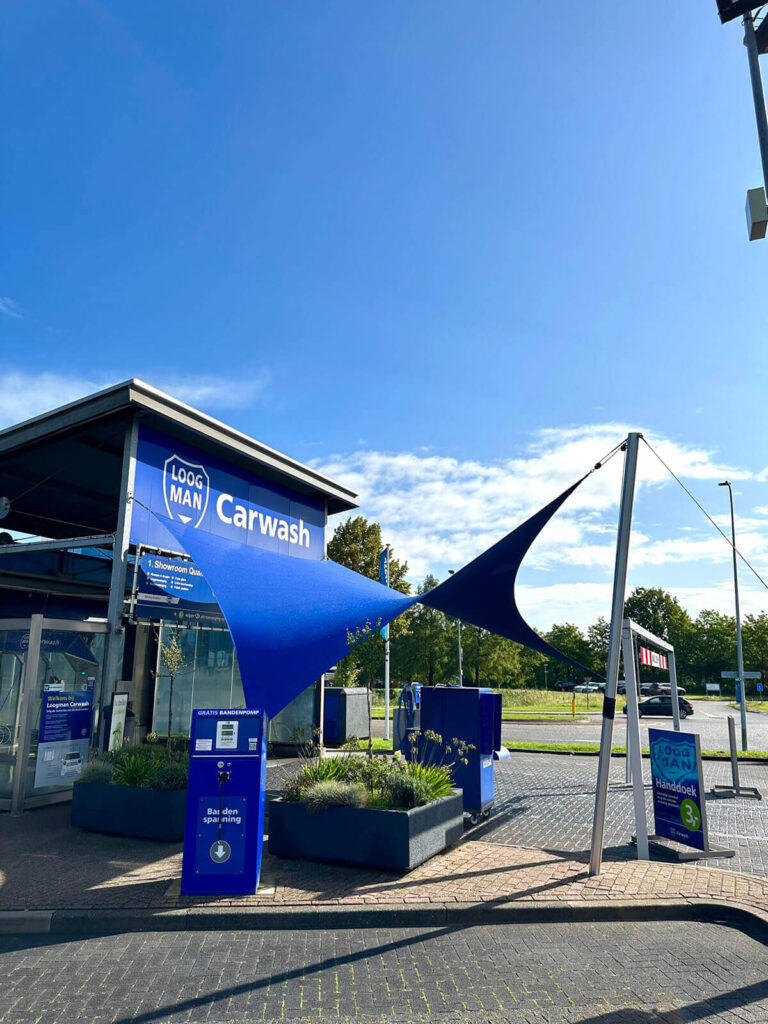 Exterior view of a carwash with blue signage and large fabric shade sails over the entrance area.