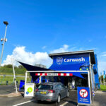 Blue carwash building with 'Carwash' sign and a gray car entering the bay under a height barrier (max 2.05 m) in daylight.