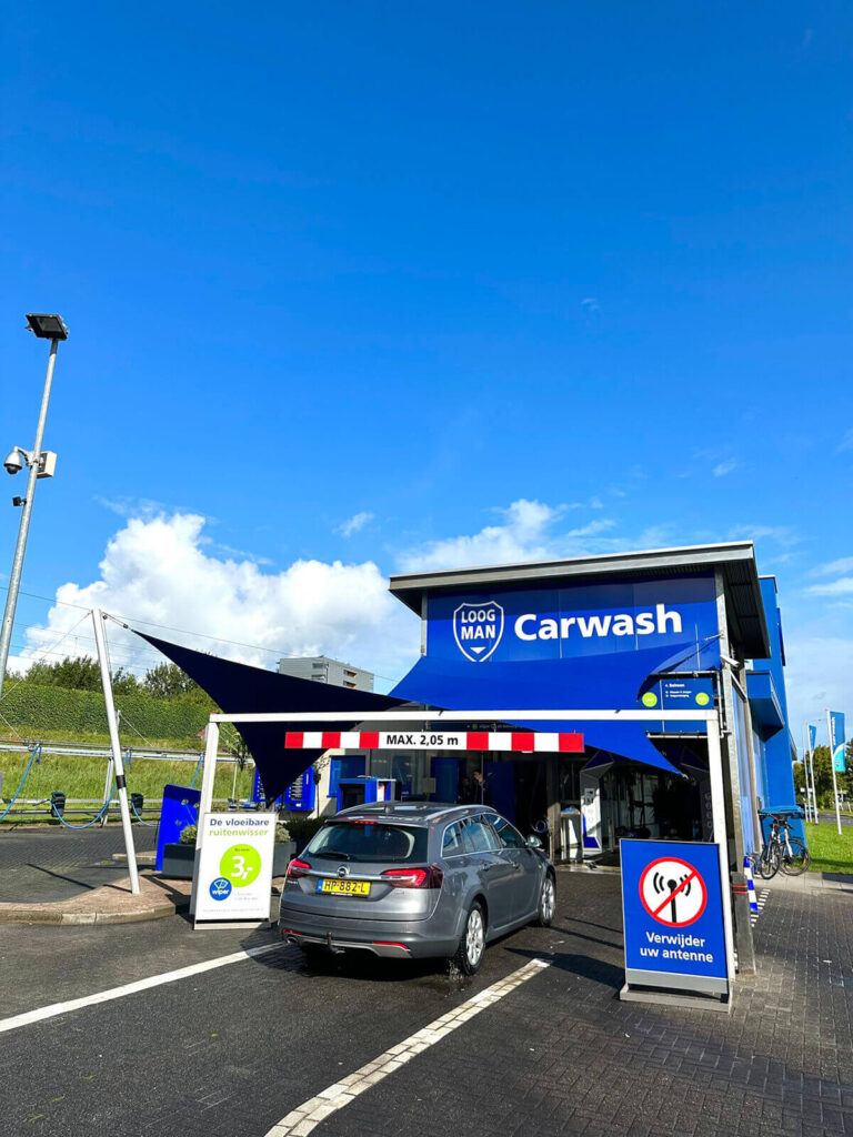 Blue carwash building with 'Carwash' sign and a gray car entering the bay under a height barrier (max 2.05 m) in daylight.