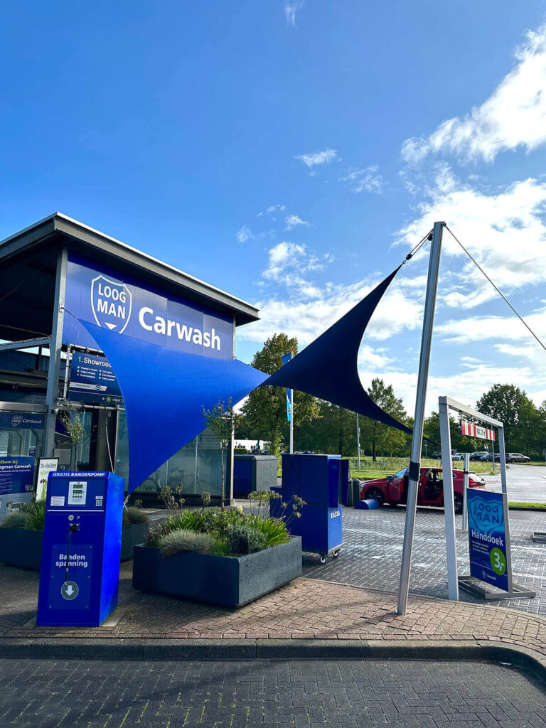 Blue car wash exterior with a large blue shade sail, blue signage reading 'Carwash', and a payment kiosk in the foreground.