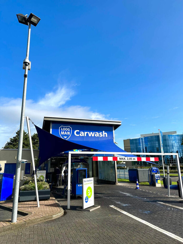 Carwash entrance with a blue canopy and a sign that reads 'Carwash' and a height limit bar marked 'MAX. 2,05 m' in front of a modern building.