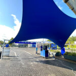 Large blue fabric shade sail stretched over an outdoor service area with blue equipment and structures in the background.