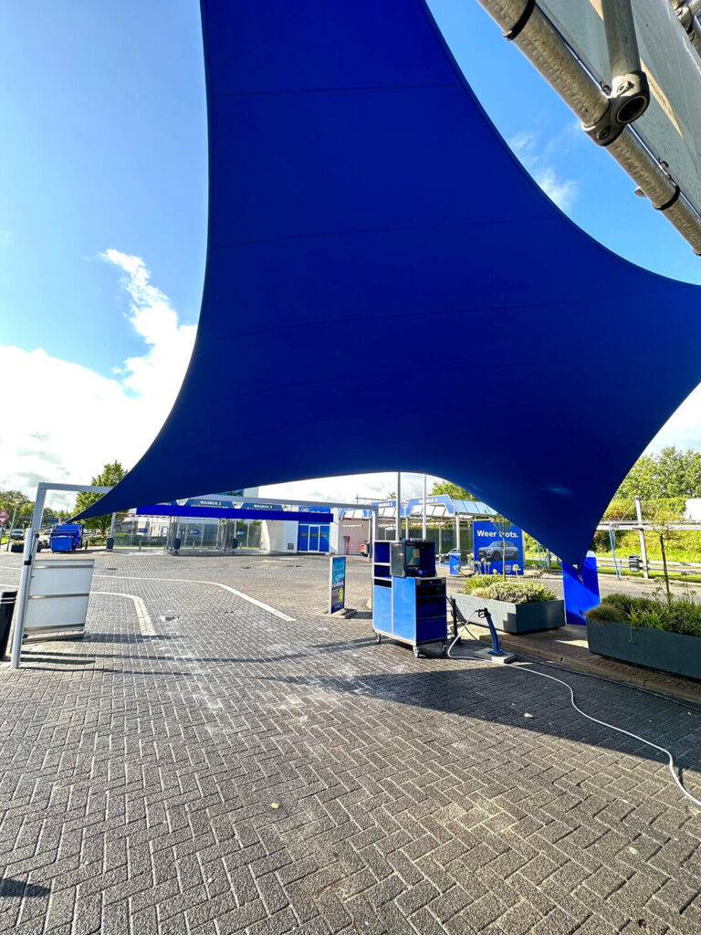 Large blue fabric shade sail stretched over an outdoor service area with blue equipment and structures in the background.