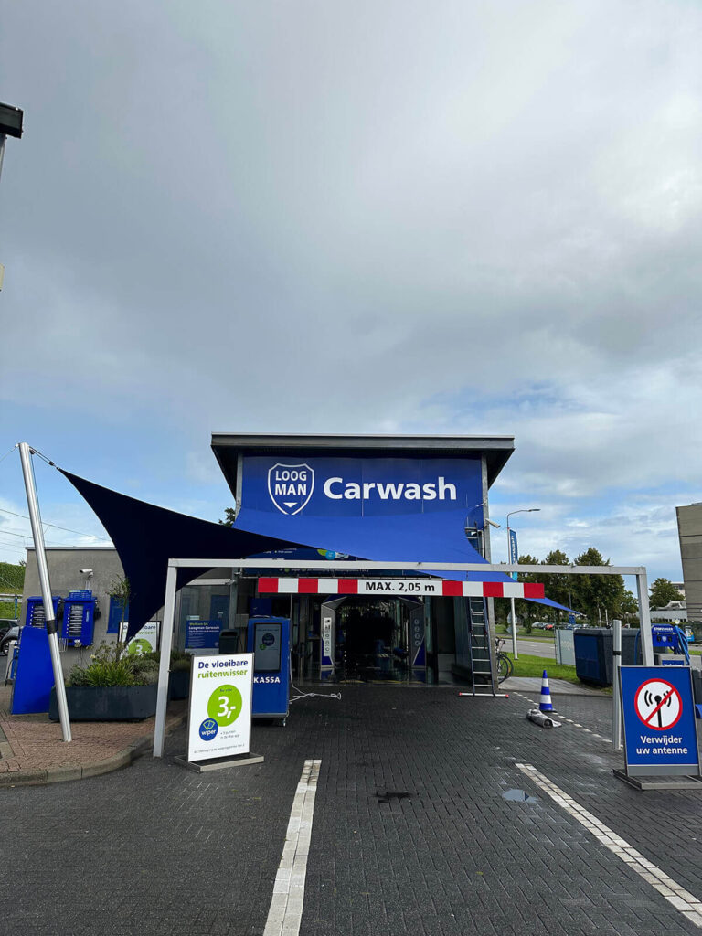Carwash entrance with a blue awning and LOOG MAN Carwash sign above, visible payment kiosk and signs at the front.