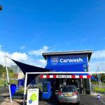 Car wash entrance with a blue canopy and a gray car ready to be washed; signs read 'Carwash' and 'KASSA' nearby.