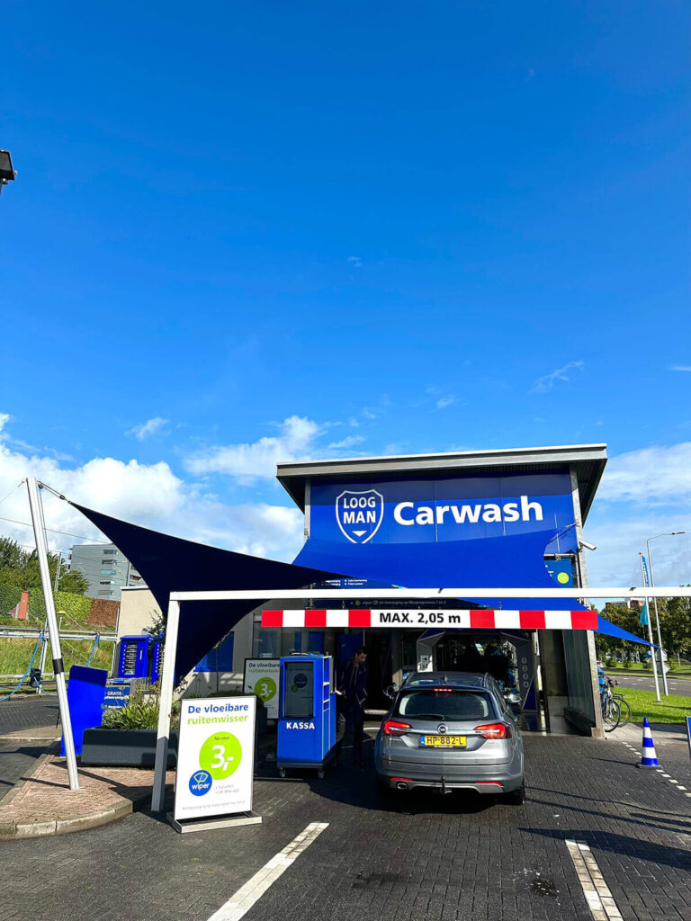 Car wash entrance with a blue canopy and a gray car ready to be washed; signs read 'Carwash' and 'KASSA' nearby.