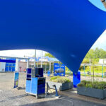 Large blue tensile shade sail over an outdoor service area with equipment cart and planters in the foreground.