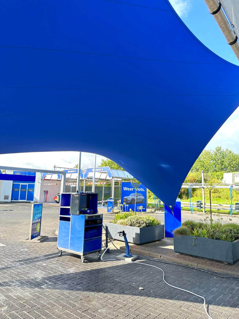 Large blue tensile shade sail over an outdoor service area with equipment cart and planters in the foreground.