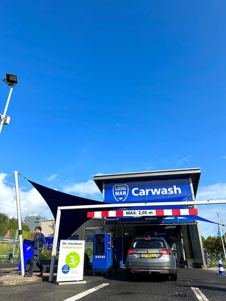 Carwash entrance with blue sign reading 'Carwash' and a gray SUV parked inside, height barrier showing 'MAX. 2,05 m' above the doorway