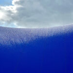 Close-up of a vivid blue curved surface with frost-like droplets, cloudy sky in the background.