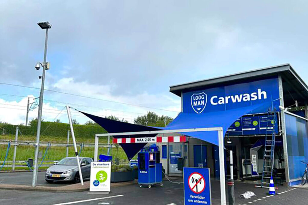 Blue car wash building with a large 'Carwash' sign and blue branding, several service kiosks, and a parked car nearby.