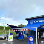 Blue car wash building with a large 'Carwash' sign and blue branding, several service kiosks, and a parked car nearby.