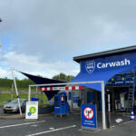 Blue car wash facility with a large 'Carwash' sign and shield logo, blue canopy sails, and a small kiosk at the entrance.