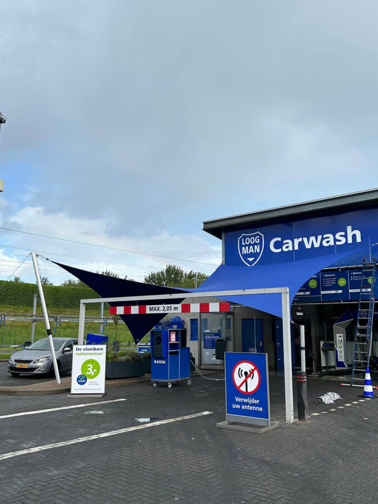 Blue car wash facility with a large 'Carwash' sign and shield logo, blue canopy sails, and a small kiosk at the entrance.
