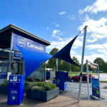 Exterior view of a blue carwash building with a large blue shade sail and a 'Carwash' sign at the entrance, payment kiosk in front.