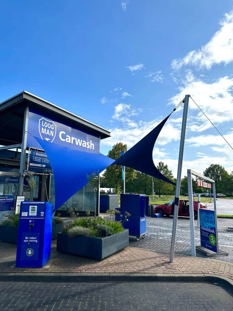 Exterior view of a blue carwash building with a large blue shade sail and a 'Carwash' sign at the entrance, payment kiosk in front.