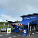 Blue carwash building with LOOG MAN logo and the word 'Carwash', bay area with signs and a barrier, and a silver car parked to the left under a cloudy sky