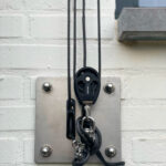 Close-up of a rope pulley system with black cables mounted on a metal plate against a white brick wall, showing hooks below.