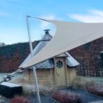 Small wooden garden shed with a curved beige shade sail stretched over it, supported by metal poles against a blue sky.