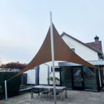 Outdoor patio with large brown fabric shade sails attached to silver poles in front of a modern white house with glass doors.