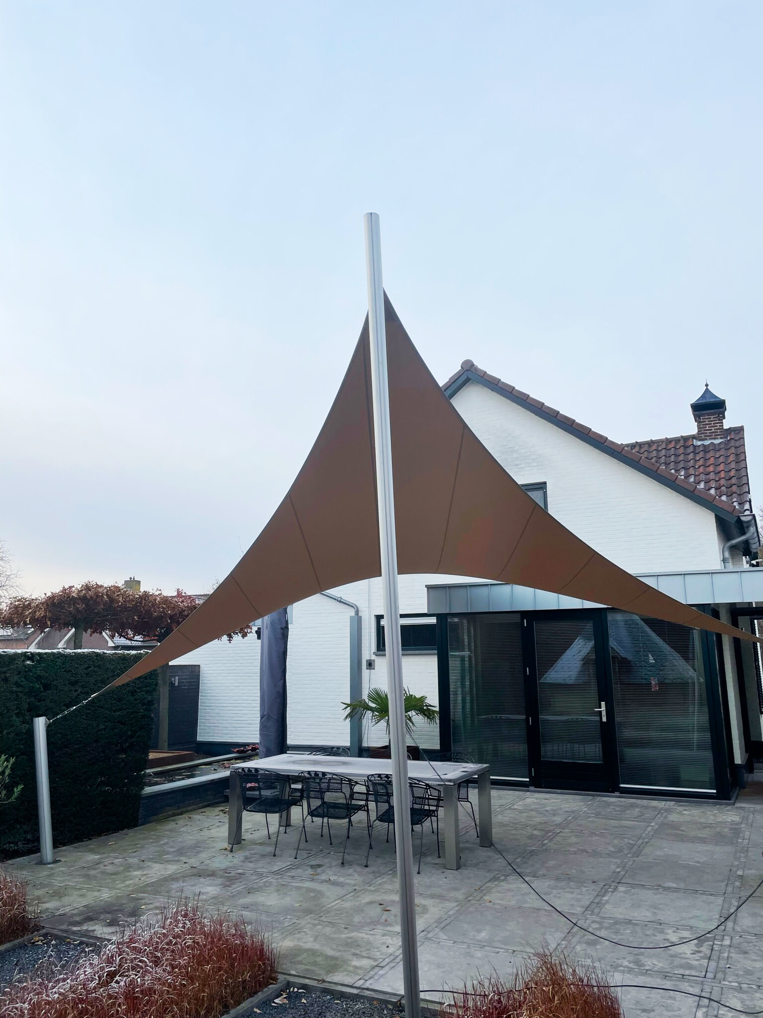 Luxe-schaduwdoek-terras-tuin-Sint_Joost-Key-Doek-design-overkapping-op-maat-key-doek-06 Outdoor patio with large brown fabric shade sails attached to silver poles in front of a modern white house with glass doors.