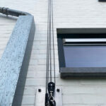 Rope-and-pulley hoist mounted on a white brick wall beside a window, with multiple cables hanging down to a metal control plate.