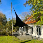 Backyard patio with a white house, black triangular shade sail, and white dining chairs under a blue sky.
