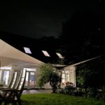 Night scene of a modern white canopy structure with lit windows and outdoor chairs on a patio at night in a garden setting.