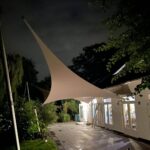 Night scene of a white building with glass doors, a large beige tensile shade sail overhead, and surrounding trees and plants at dusk.