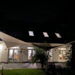 Night exterior of a white brick building with illuminated interior through large glass doors and sails stretched overhead.