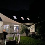 Nighttime backyard with a white fabric sail shade over a patio; people are visible inside the house through glass doors.