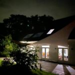 Night view of a modern building with a curved white canopy and illuminated glass doors, surrounded by greenery.