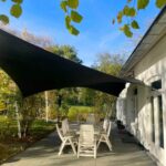 Outdoor patio with white plastic chairs around a table beneath a large black shade sail; garden and blue sky in the background.