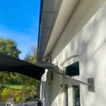 White brick house exterior with a black patio shade sail over an outdoor seating area of white chairs and tables.