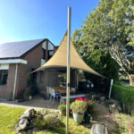 Backyard patio by a brick house with solar panels, a tall beige shade sail, and a wooden dining table with chairs.