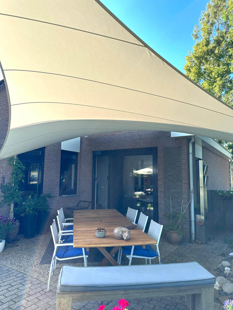 Outdoor dining area under a large cream shade sail attached to a brick house; wooden table with white chairs and blue cushions, potted plants nearby.