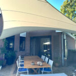 Outdoor dining area under a large beige shade sail over a wooden table with white chairs and blue cushions, brick house in background.