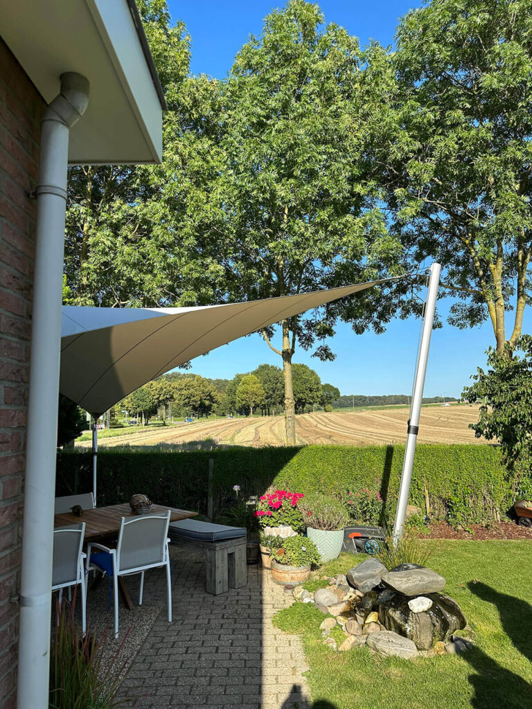 Backyard patio with a white shade sail, a brick wall on the left, and a table with chairs, overlooking a field and trees beyond.