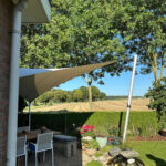 Outdoor patio with cream shade sails, brick wall on left, table and chairs, potted plants, and a manicured hedge; field beyond under blue sky.