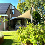 Brick house with solar panels, a beige shade sail over a patio, and a hanging basket of green plants in a sunny backyard