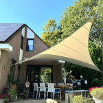 Backyard patio of a brick house with a large beige triangular shade sail over a dining table and white chairs; a dog stands nearby.